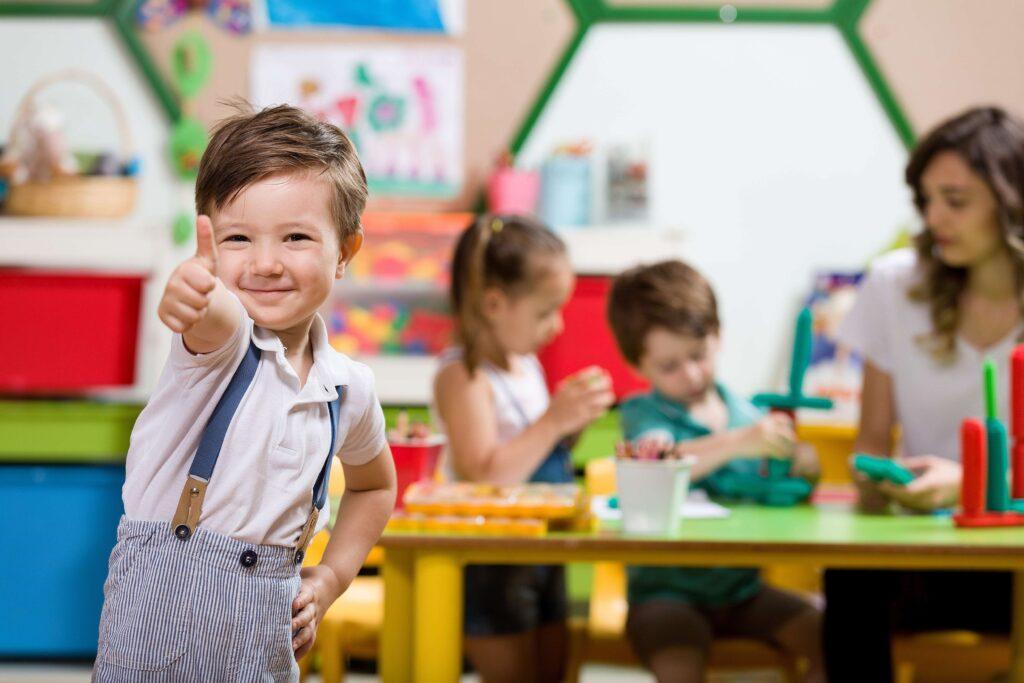 Ein Junge zeigt einen Daumen hoch in die Kamera und lächelt. Dahinter sitzen Kinder an einem Tisch.