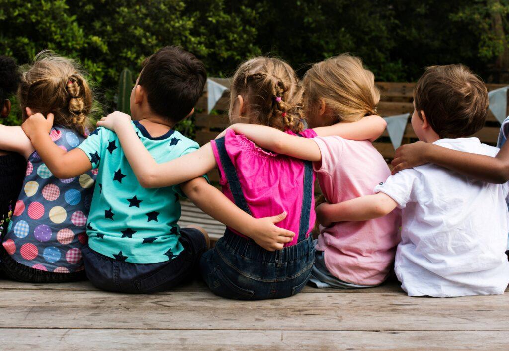 Kinder sitzen mit dem Rücken gewandt auf dem Boden und haben ihre Arme über ihre Schultern gelegt.