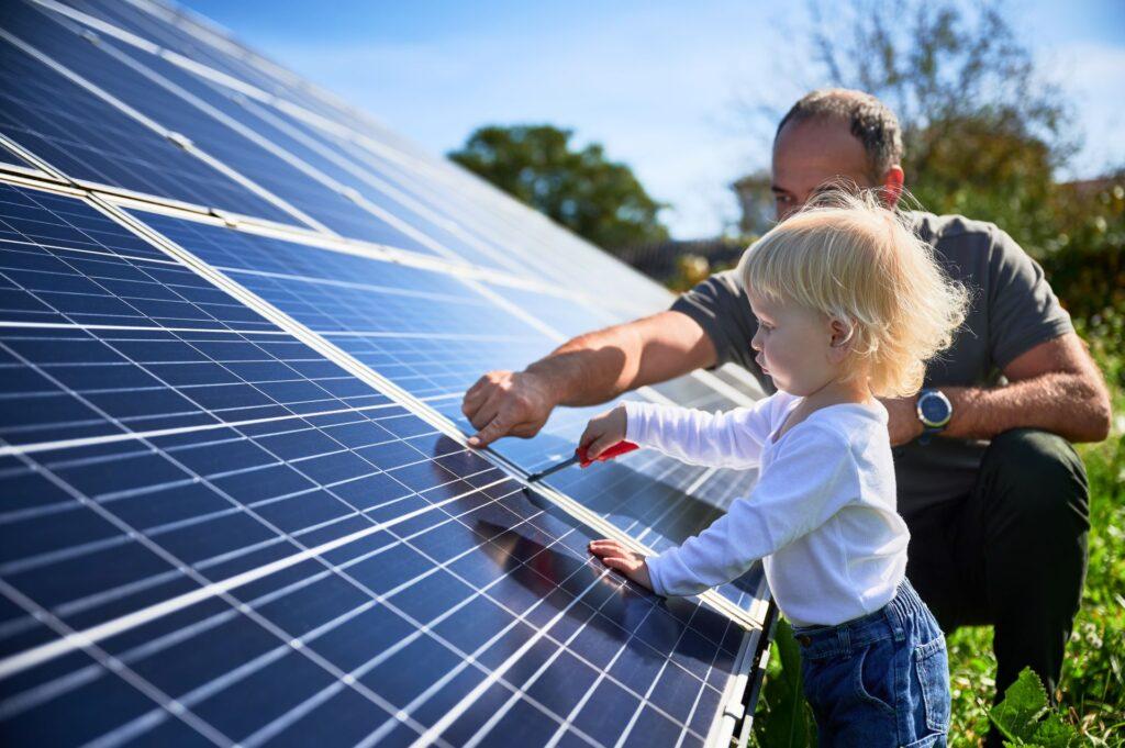 Kleines Kind und Vater arbeiten gemeinsam an einer Solaranlage im Garten. Das Bild zeigt die familiäre Zusammenarbeit und den Einsatz erneuerbarer Energien durch Solarenergie.