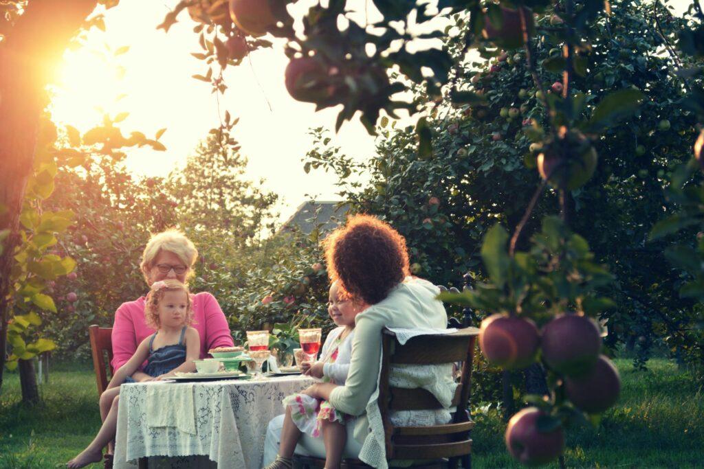 Familientreffen im Obstgarten bei Sonnenuntergang: Ältere Frau und Mutter mit zwei Kindern am Tisch im Grünen