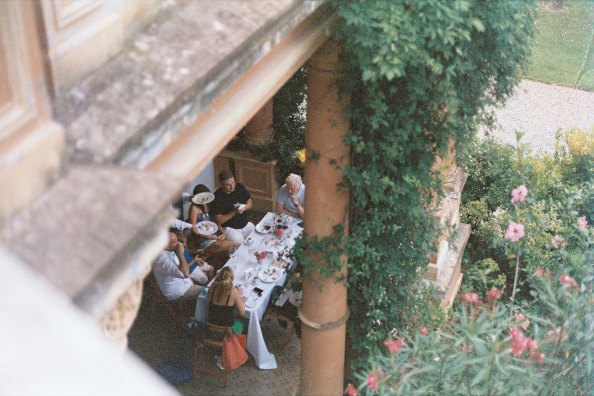 Gartenfeier in einem mediterranen Innenhof, mehrere Personen sitzen um einen gedeckten Tisch, umgeben von grünen Pflanzen und Blumen